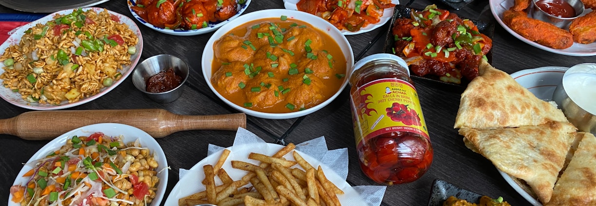 A diverse spread of Indian cuisine on a dark table, including dishes with vibrant colors and various textures. There are plates of bright red chicken dishes, golden fried potatoes with green garnish, and creamy curries. Accompanying these are crispy flatbreads, pickles, samosas, and a selection of dipping sauces. Also visible are rice dishes with vegetables and spices, along with a jar of spicy chili pickles.