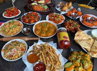 A diverse spread of Indian cuisine on a dark table, including dishes with vibrant colors and various textures. There are plates of bright red chicken dishes, golden fried potatoes with green garnish, and creamy curries. Accompanying these are crispy flatbreads, pickles, samosas, and a selection of dipping sauces. Also visible are rice dishes with vegetables and spices, along with a jar of spicy chili pickles.
